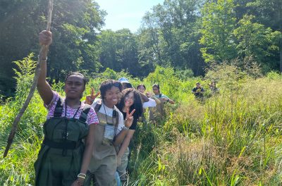 A group of students walk in a line in tall grass.